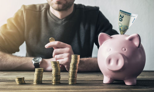 Mann stapelt Münzen auf, daneben steht ein Sparschwein mit einem 20-Euro-Schein.